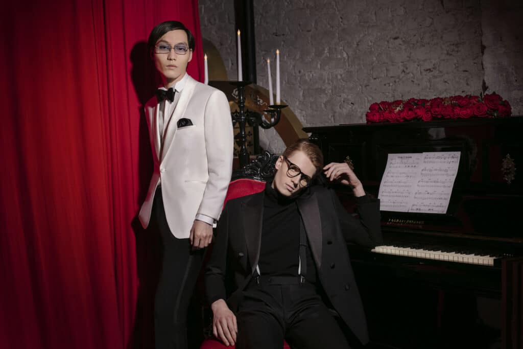 Zwei elegant gekleidete Männer, einer im weißen Smoking vor rotem Samtvorhang stehend, der andere entspannt am Klavier mit Rosen.